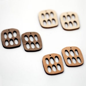 Puede incluir: Pendientes de madera en varios tonos de marrón y beige. Cada pendiente tiene forma rectangular con esquinas redondeadas y seis recortes ovalados. Un pequeño agujero en la parte superior para colgar.
