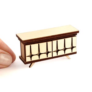 May include: A miniature wooden sideboard featuring a light beige top and base with dark brown trim. The two front cabinet doors display a repeating cutout pattern of rounded shapes. The piece is supported by four small, angled wooden legs.