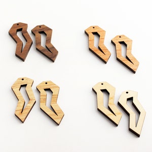 May include: Four pairs of wooden earrings in various shades of brown and tan. Each earring has a geometric, open design with a small hole at the top for hanging. The earrings are displayed on a white surface.