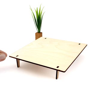 May include: A small, unfinished wooden table with four legs and a square top. The table is made of light-colored wood and has small square cutouts at each corner. A small wooden planter with a green plant is in the background.