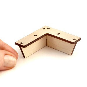 May include: A small, L-shaped wooden corner piece with a light wood grain finish and dark brown edges. The piece has several small square holes and is designed to fit together at a 90-degree angle. The corner piece is shown against a white background.