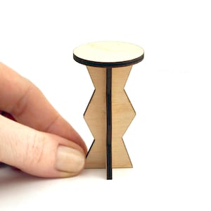 May include: A small, unfinished wooden table with a round top and a zig-zag patterned base. The table is made of light-colored wood with dark brown edges and accents. The table is approximately 2 inches tall.