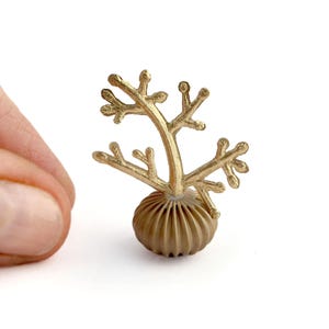May include: A small, gold-colored decorative object resembling a stylized coral or plant. It features a textured, ribbed base and branching, twig-like structures extending upwards. The object is held against a white background.