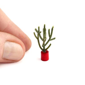 May include: A miniature, green, plant-like sculpture in a small, red, cylindrical pot. The plant has multiple, thin, branch-like structures. The image is set against a white background.