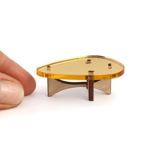 Könnte beinhalten: Miniatur-Couchtisch mit einer transparenten, bernsteinfarbenen Platte und Holzbeinen. Der Tisch hat ein modernes Design aus der Mitte des Jahrhunderts. Der Tisch ist etwa 5 cm lang und 2,5 cm breit.