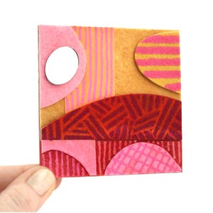 Puede incluir: Una pequeña obra de arte abstracta cuadrada con una mezcla de texturas y colores. Presenta formas rosas, naranjas y rojas, incluyendo un círculo, rayas y patrones geométricos. La pieza es sostenida por una mano sobre un fondo blanco.