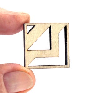 May include: A small, square wooden cutout with geometric shapes. The cutout features a triangle and an L-shaped design, with the negative space creating the shapes. The wood has a natural, light color with dark edges.