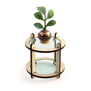 May include: A miniature wooden side table with two clear glass shelves. A small copper-colored sphere with a green plant sits on the top shelf. The table is made of light-colored wood and has a round design.