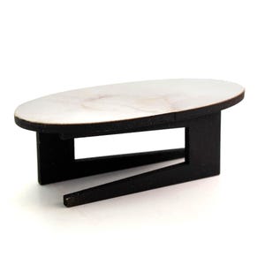 Puede incluir: Una mesa de madera en miniatura con base negra y tapa ovalada de color claro. La mesa tiene un diseño moderno, adecuada para casas de muñecas o proyectos de manualidades. La mesa mide unos 10 cm de largo.