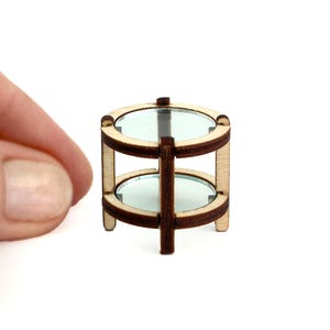 May include: Miniature two-tiered table with clear glass tops and a light wood frame. The table has a dark brown central support and is designed for a dollhouse or similar craft project. The table is approximately 1.5 inches tall.