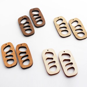 Puede incluir: Pendientes de madera en tonos marrones y beige. Cada par tiene forma rectangular con tres cortes ovalados y un pequeño agujero para colgar. Los pendientes están hechos de madera natural.