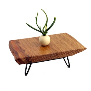 May include: A rectangular wooden table with a natural live edge and a smooth grain finish, supported by four thin black metal hairpin legs. A small, round, off-white decorative object with green stems sits centered on the wooden surface.