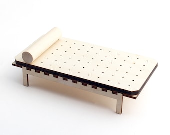 May include: A miniature wooden bed with a light-colored finish. The bed features a rectangular base with a perforated surface and a cylindrical headrest. The bed is supported by four legs and is designed for decorative purposes.