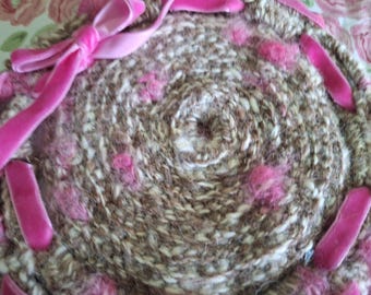 Trivet crocheted in beige and brown handspun yarn with pink accents.  A pink velvet ribbon is laced through open stitching and tied in a bow
