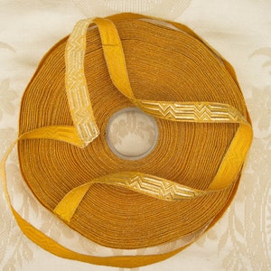 May include: A roll of mustard yellow ribbon featuring a silver geometric design. The ribbon is coiled, with a section unravelling to display the pattern. The background is a cream-coloured fabric with a delicate floral motif.