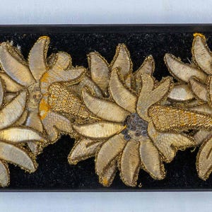 Könnte beinhalten: Nahaufnahme einer schwarzen Samtborte mit goldenen gestickten Edelweißblüten. Die Blüten sind in einem sich wiederholenden Muster angeordnet.