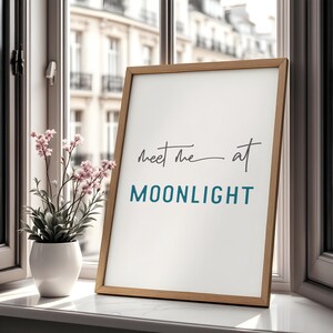 May include: A framed print with a white background and a wooden frame. The print features the text "Meet me at MOONLIGHT" in black and blue lettering.