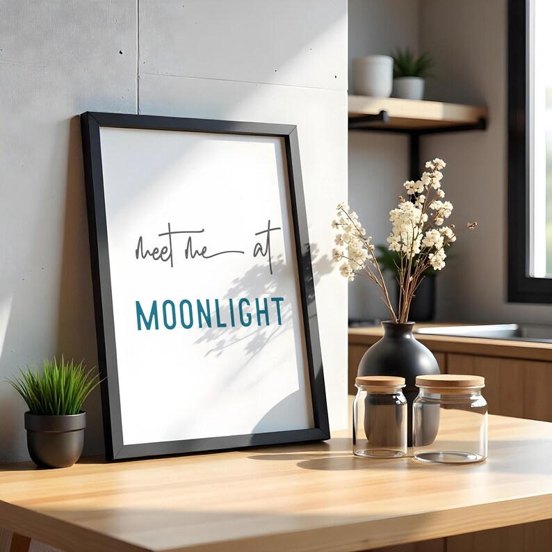 May include: A black framed print with the text "Meet me at MOONLIGHT" in white and teal lettering. The print is sitting on a wooden table with a vase of white flowers and two glass jars with wooden lids.