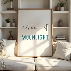May include: A white framed print with the text "Meet me at MOONLIGHT" in black and teal lettering. The print is hanging on a white wall above a white couch with pillows.