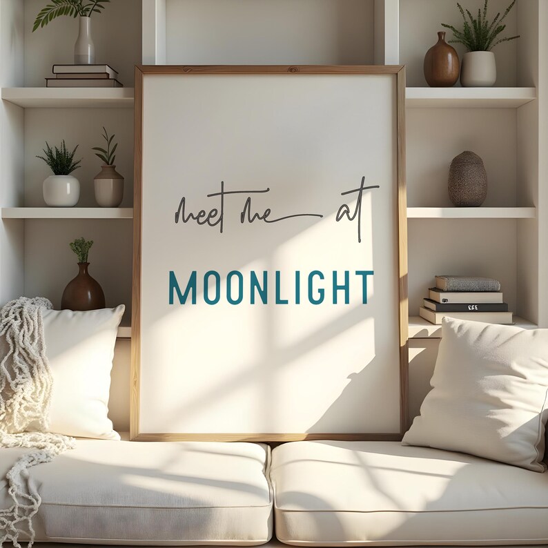 May include: A white framed print with the text "Meet me at MOONLIGHT" in black and teal lettering. The print is hanging on a white wall above a white couch with pillows.