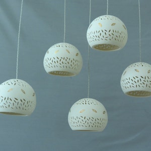 Peut inclure: Cinq lampes suspendues en c&eacute;ramique blanche avec des accents de feuilles d'or et un motif d&eacute;coup&eacute; pendent &agrave; des cordons blancs sur un fond bleu clair.
