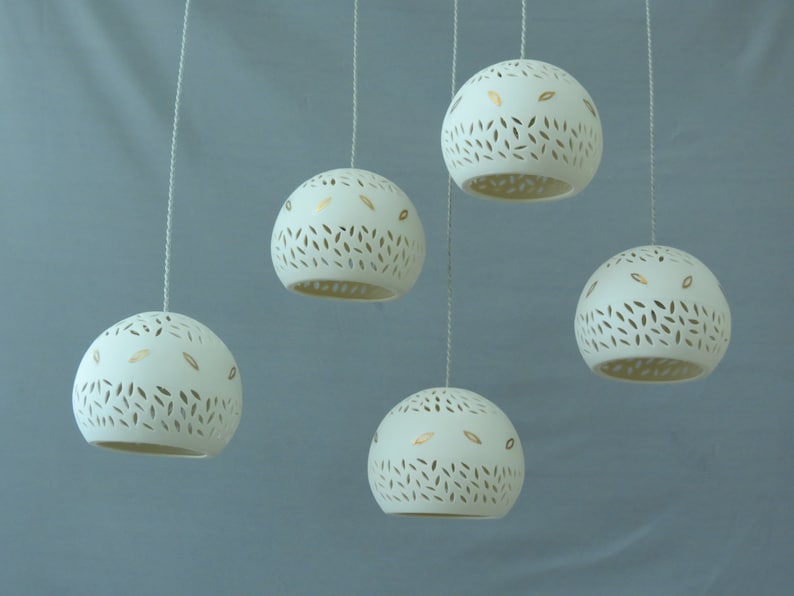 Peut inclure: Cinq lampes suspendues en c&eacute;ramique blanche avec des accents de feuilles d'or et un motif d&eacute;coup&eacute; pendent &agrave; des cordons blancs sur un fond bleu clair.