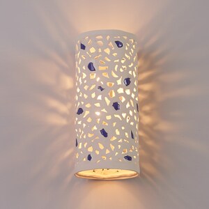 May include: A white ceramic wall sconce with a cylindrical shape. The sconce has a perforated design with irregular cutouts and is adorned with blue accents. The light shines through the cutouts, creating a pattern on the wall.