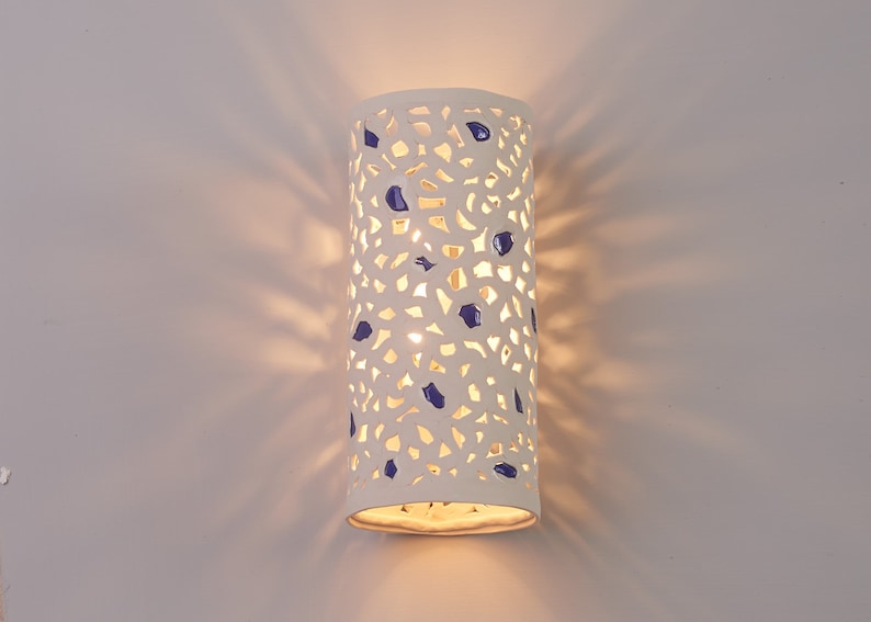 May include: A white ceramic wall sconce with a cylindrical shape. The sconce has a perforated design with irregular cutouts and is adorned with blue accents. The light shines through the cutouts, creating a pattern on the wall.