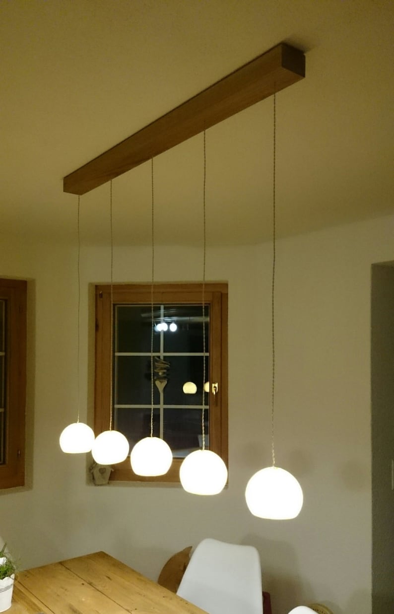 May include: A wooden ceiling fixture with five white globe-shaped pendant lights hanging from it. The lights are hanging over a wooden table with a white chair in the foreground.