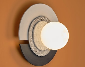 Puede incluir: Aplique de pared moderno con una luz esférica. El aplique presenta un diseño en capas con elementos semicirculares gris claro, blanco roto texturizado y gris oscuro. La luz es una esfera brillante y luminosa.