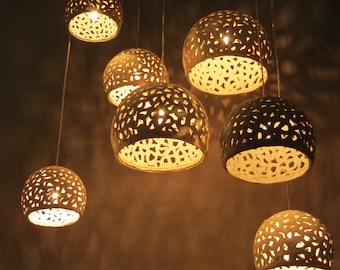 May include: A cluster of seven pendant lights with a spherical design. Each light has a perforated pattern, allowing the warm light from the bulbs inside to shine through. The lights hang from thin cords against a dark background.