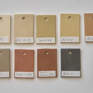 Op de afbeelding: Tien kleine keramische tegels in verschillende kleuren, waaronder wit, gebroken wit, gespikkeld, geel, bruin, grijs, terracotta, donker terracotta en zwart. Elke tegel heeft een klein gaatje in het midden en een label met de naam van de kleur erop.
