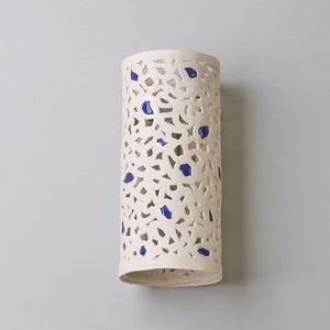 May include: A cylindrical, off-white ceramic wall sconce with a cut-out floral design and scattered blue accents. The sconce is approximately 12 inches tall and has a smooth, matte finish. It is designed to hold a light source.