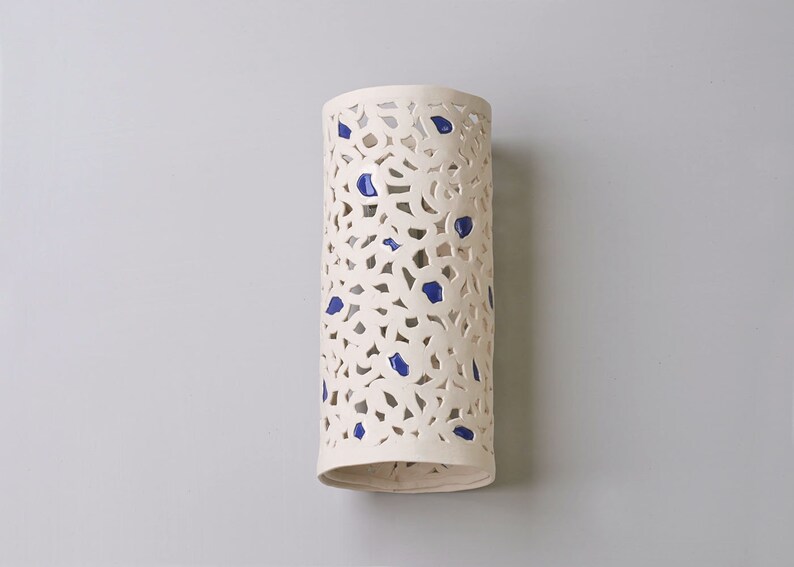 May include: A cylindrical, off-white ceramic wall sconce with a cut-out floral design and scattered blue accents. The sconce is approximately 12 inches tall and has a smooth, matte finish. It is designed to hold a light source.