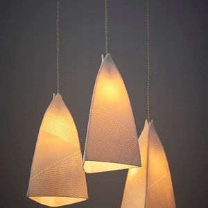 May include: Three white ceramic pendant lights with a wavy pattern hang from the ceiling. The lights are illuminated and cast a warm glow.
