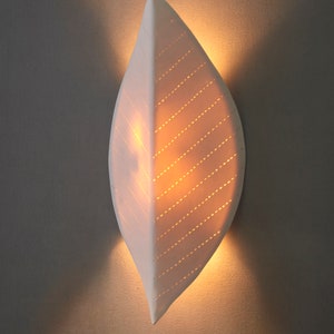 May include: A white ceramic wall sconce shaped like a leaf with a light inside. The leaf has a pattern of small holes that allow light to shine through.