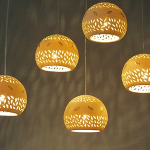 Peut inclure: Cinq luminaires suspendus avec un abat-jour en c&eacute;ramique blanche. Chaque abat-jour pr&eacute;sente un motif d&eacute;coup&eacute; de petites feuilles. Les lumi&egrave;res sont allum&eacute;es et brillent.