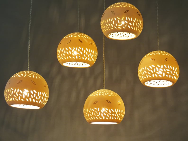 Peut inclure: Cinq luminaires suspendus avec un abat-jour en c&eacute;ramique blanche. Chaque abat-jour pr&eacute;sente un motif d&eacute;coup&eacute; de petites feuilles. Les lumi&egrave;res sont allum&eacute;es et brillent.