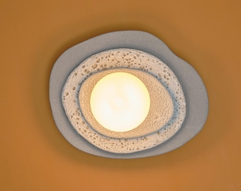Puede incluir: Una moderna lámpara de techo abstracta. La luz tiene una bombilla central redonda y brillante, rodeada por tres capas concéntricas de forma orgánica. La capa exterior es de un gris azulado apagado, la capa intermedia es blanca texturizada y la capa interior es una luz cálida.