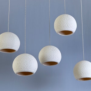 May include: Five white ceramic pendant lights with a textured surface hang from the ceiling. The lights are round and have a hole in the bottom.