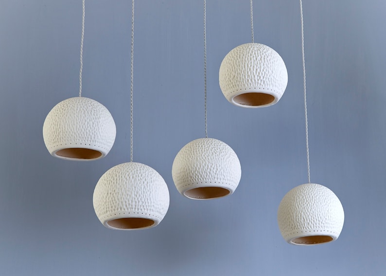 May include: Five white ceramic pendant lights with a textured surface hang from the ceiling. The lights are round and have a hole in the bottom.