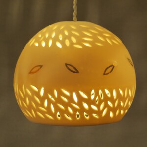 Peut inclure: Un luminaire suspendu en c&eacute;ramique blanche de forme ronde avec des motifs d&eacute;coup&eacute;s complexes. La lumi&egrave;re est allum&eacute;e, projetant une lueur chaude &agrave; travers les d&eacute;coupes.