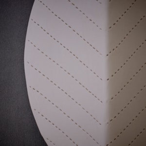 May include: A white circular piece of fabric with a pattern of diagonal dashed lines. The fabric is stitched together with a decorative stitch.