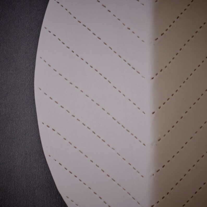 May include: A white circular piece of fabric with a pattern of diagonal dashed lines. The fabric is stitched together with a decorative stitch.