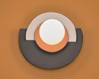 Puede incluir: Aplique de pared moderno con un diseño geométrico. La lámpara presenta un globo blanco y brillante en el centro, rodeado por un círculo de color terracota, un semicírculo gris claro y un semicírculo gris oscuro. Montado en una pared naranja cálida.