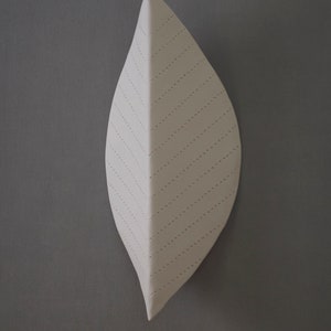 May include: A white ceramic leaf wall sculpture with a stitched design. The leaf is shaped like a teardrop and has a textured surface.