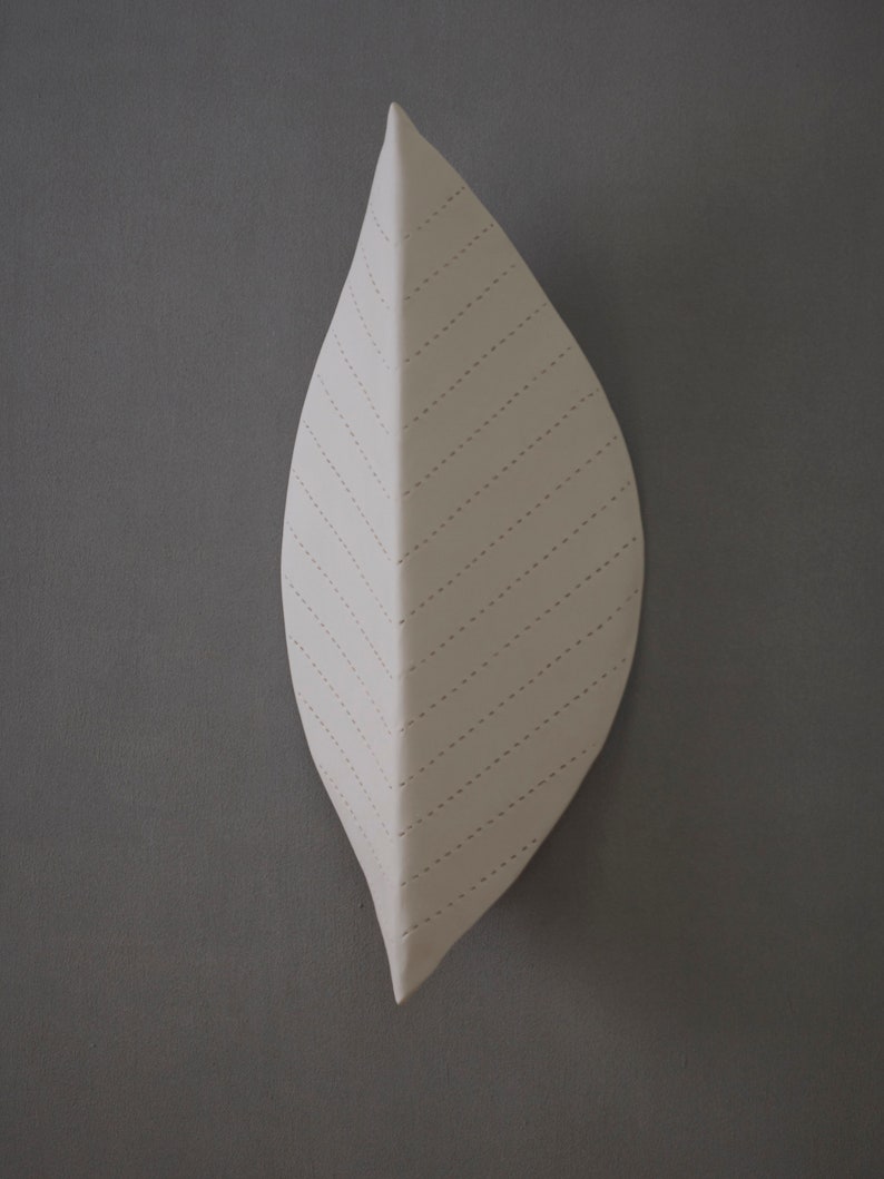 May include: A white ceramic leaf wall sculpture with a stitched design. The leaf is shaped like a teardrop and has a textured surface.
