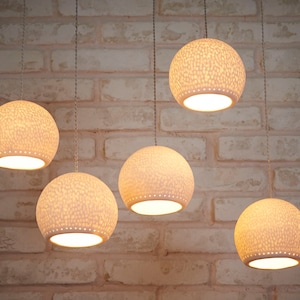 May include: Five white, round, hanging light fixtures with a textured surface. The lights are hanging from a brick wall.