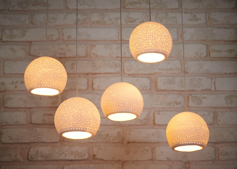 May include: Five white, round, hanging light fixtures with a textured surface. The lights are hanging from a brick wall.