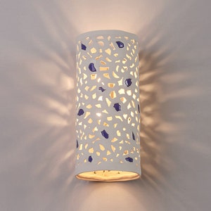 May include: A white ceramic wall sconce with a cylindrical shape. The sconce features a cut-out design with blue accents. The light shines through the cut-outs, creating a pattern on the wall. The sconce is mounted on a white wall.